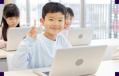 教室でノートパソコンに向かい、ピースサインをして笑顔を見せる子ども